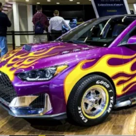 A custom purple sports car with yellow and orange flame decals, displayed at an indoor auto show with people in the background.