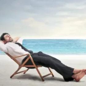 A businessman in a suit relaxing on a wooden chair at the beach, leaning back with his hands behind his head.